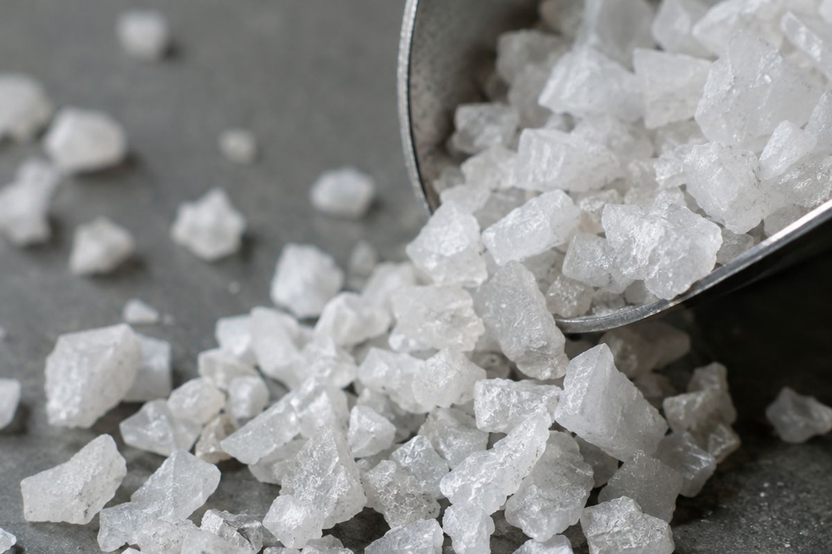 Coarse white silica granules spilling from a stainless steel scoop onto a laboratory surface